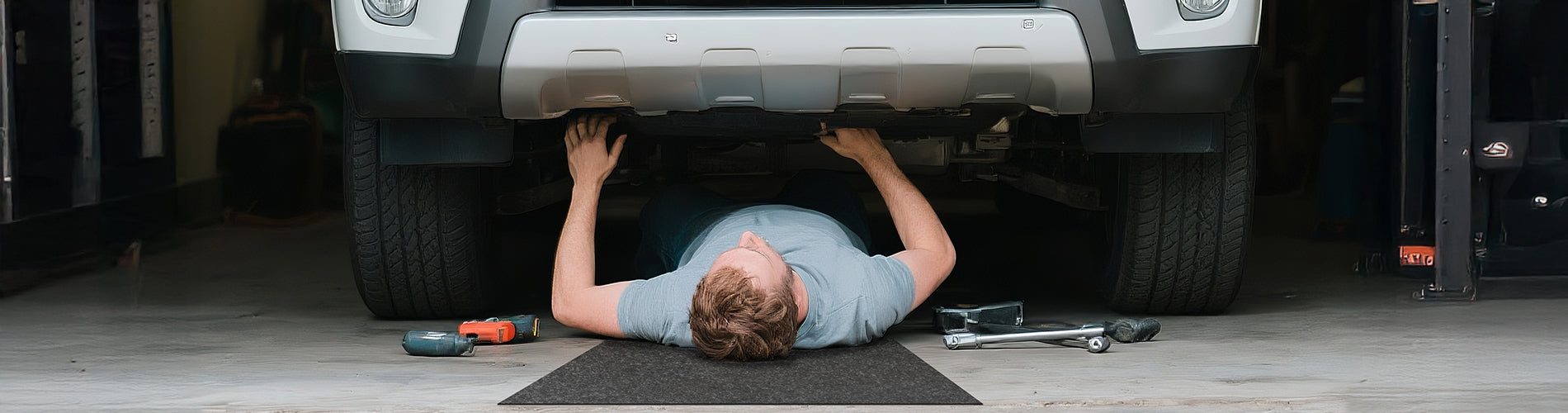 Accessory image for the WELKIN Garage Floor Mat (A1204) showing a person lying on the mat under an SUV performing car maintenance, highlighting its use as a comfortable creeper pad for vehicle repair.