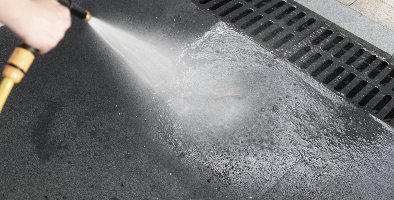 Accessory image for WELKIN Garage Floor Mat (A1204) showing a hand using a yellow hose nozzle to spray water and clean the dark floor mat, emphasizing the mat's easy-to-clean and washable feature.