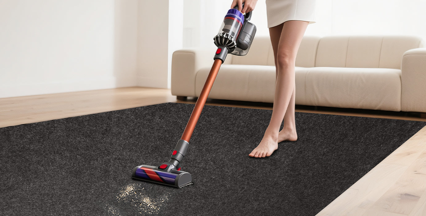 Accessory image for WELKIN Garage Floor Mat (A1204) showing a person using a cordless vacuum cleaner on the dark gray mat in a living room setting, highlighting its ease of maintenance and vacuum-friendly texture.