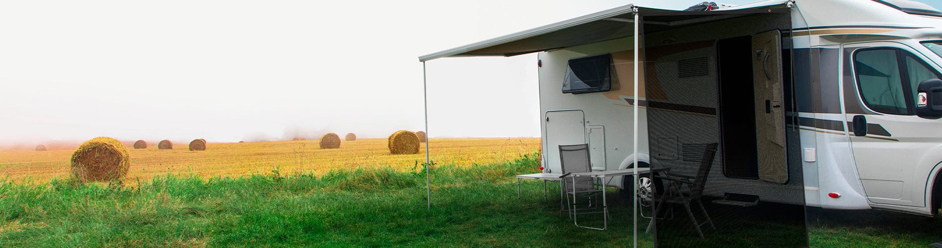 Homepage banner image showing a white RV parked in a grassy field with hay bales in the background, featuring the awning extended over a dining set for outdoor camping leisure.